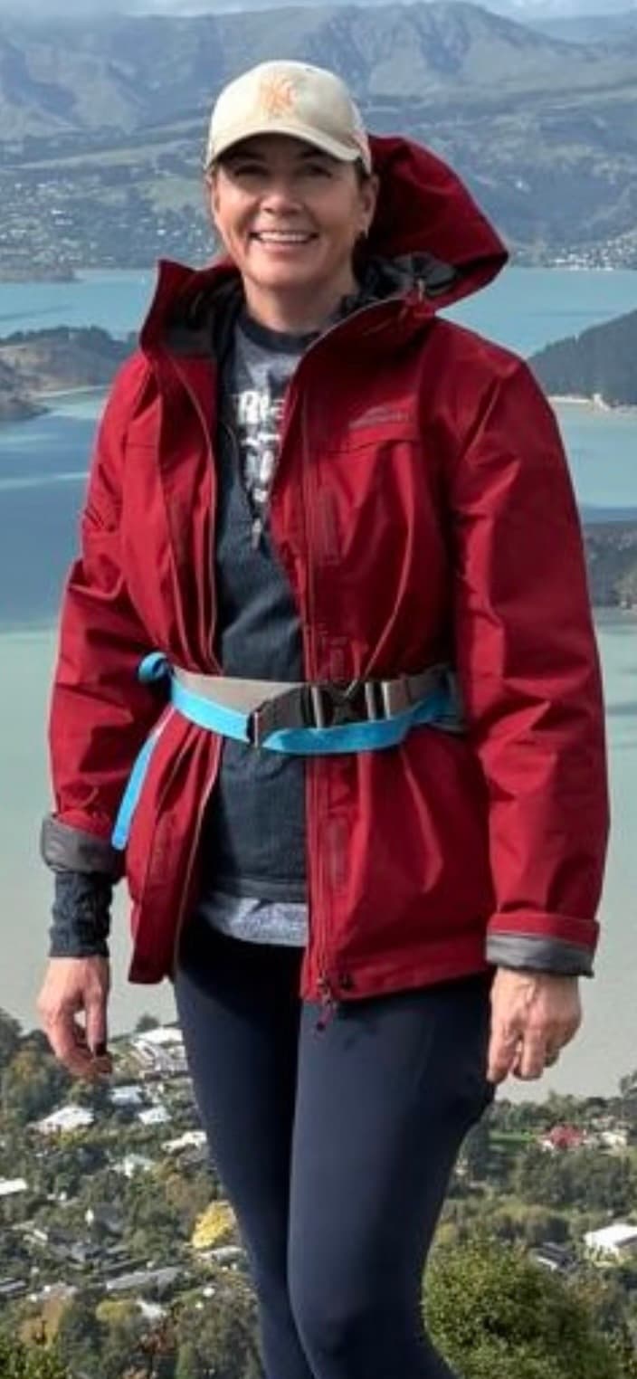 Stephanie Sisson, founder of Your Person NZ, smiling in a red jacket and cap on a hillside above Lyttelton Harbour