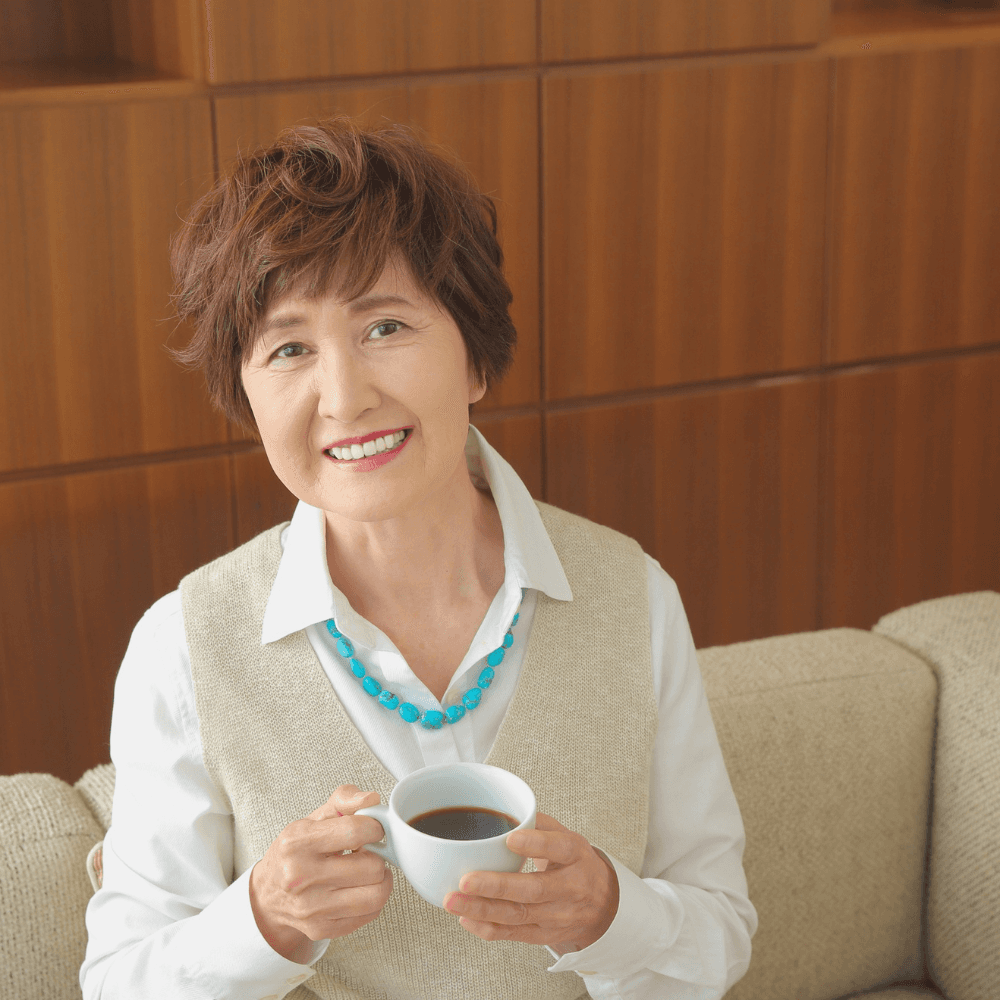 A smiling older woman in a cream knitted vest sitting on a sofa, holding a cup of coffee
